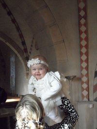 Kleinkind mit weißem Stirnband und Taufkleid in den hoch erbobenen Armen der erwacnsenen Patin nach der Taufe in St. Katharinen, Schönemoor.