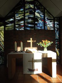 Altar des St. Johannes Hauses von Sonnenstrahlen erhellt, Blumenschmuck und brennende Kerzen, im Hintergrund das Buntglasfenster, weißes Parament für das Christusfest Ostern.