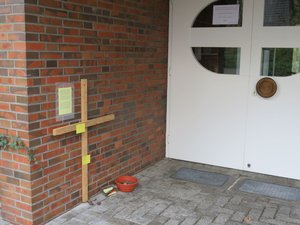 Holzkreuz aus hellem Holz vor der Klinkerwand im Eingang des St. Johannes Hauses Hasport-Annenheide mit einer Anleitung, Sorgen und Ängste am Kreuz auf einem Zettel abzuladen. 