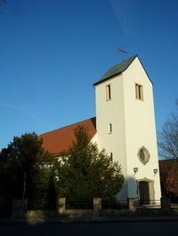 Die weißen Wänder der St. Hedwigkirche in Ganderkesee vor blauem Himmel, rote Ziegel auf dem Dach, Kreuz auf dem Kirchturm.