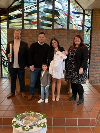 Familienbild nach der Taufe, Säugling im Taufkleind auf dem Arm der Mutter, der Bruder der Kleinen vor den Eltern, rechts und links Patin und Pate mit großer Taufkerze. 