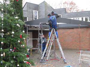 Handwerker errichten vor dem Eingang zum Johannes Haus den Rahmen für ein Glasdach - am linken Bildrarnd ist der geschmückte Weihnachtsbaum zu erkennen.