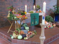 Erntedankschmuck verdeckt den Altartisch, im Vordergrund die Taufkerze auf dem gedrechselten Ständer, verschiedene Kohlsorten, Obst im Korb Möhren und Brot auf dem Tisch.