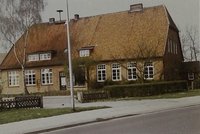Schulgebäude hinter Zaun und Hecke, moderne Straßenlaterne, Asphaltstraße im Vordergrund