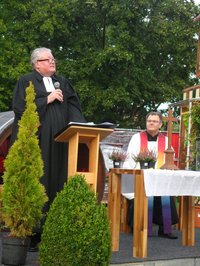 Pfarrer Michael Stuhlken mit Mikrofon hinter einem Pult stehend, Pfarrer Guido Wachtel hinter einem Altar sitzend, Blumendekoration.