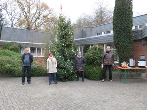 Weihnachtsbaum mit Kugeln und Lichterketten, rote Weihnachtsmann Mütze auf der Baumspitze, vier Menschen mit Abstand vor dem Baum, im Hintergrund das St. Johannes Haus in Hasport-Annenheide.