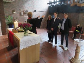Pfarrerin Ulrike Klank mit Mundschutz am Altar mit Blumenschmuck und berennenden Kerzen; ihr gegenüber der Konfirmand Paul Behlke mit seinen Eltern. 