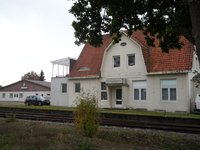 Bahnhofsgebäude Annenheide, Schützenhalle im Hintergrund, Schienenstrang vorn unter Bäumen.
