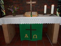 Hölzerner Altartisch mit Spitzendecke, zwei brennende Kerzen, Bibel vor einem Bronzekreuz